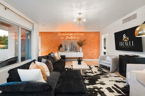 A modern living room with a black sofa, white armchairs, and a cowhide rug. A neon sign reads 'It's always sunny in Scottsdale.' - NEW|Iconic Granada Grotto|Pool & Hottub| Sleeps 12 (Scottsdale)