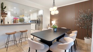 Une cuisine moderne avec des meubles blancs, un réfrigérateur en acier inoxydable et un coin repas comprenant une table foncée et des chaises blanches.