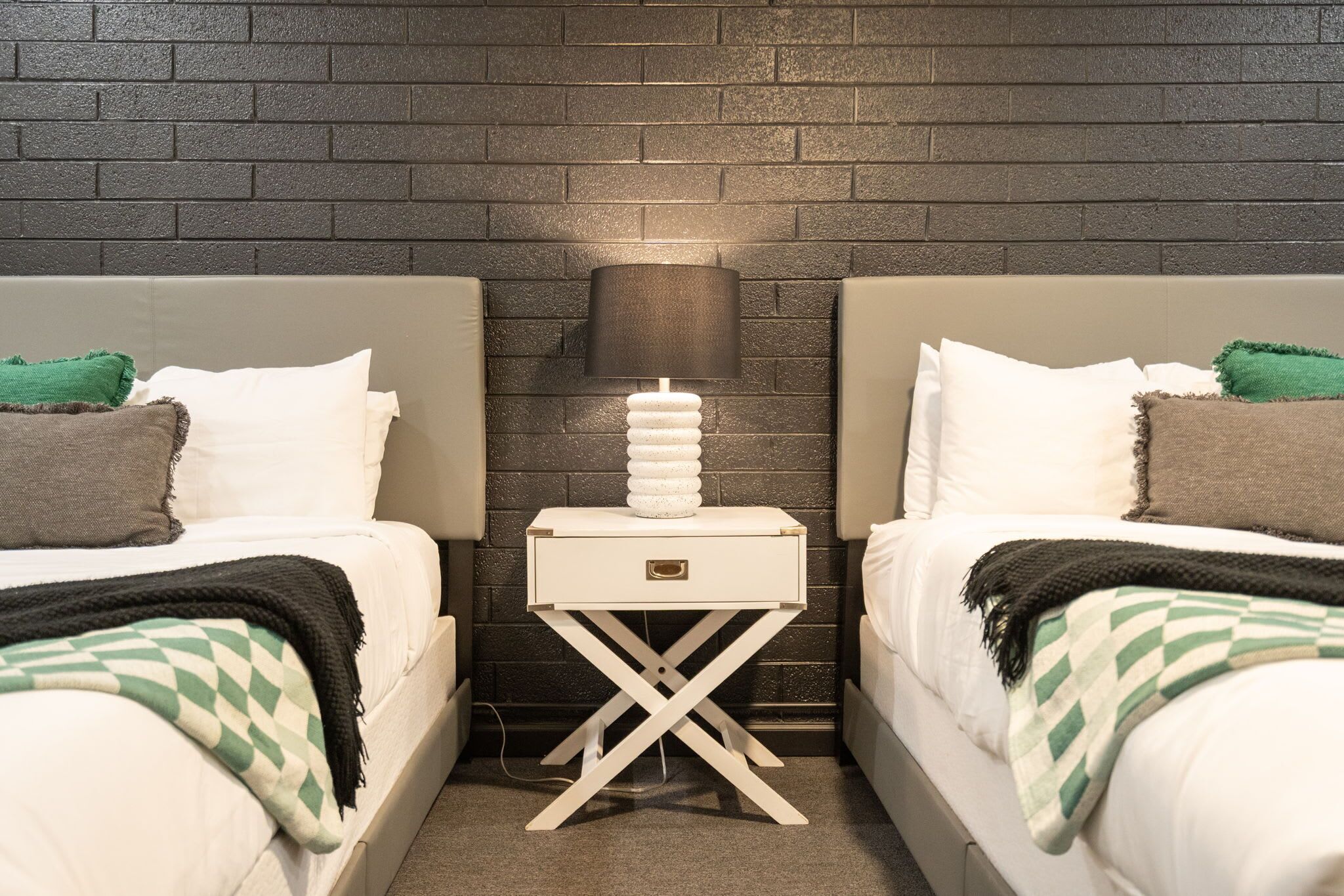 A hotel room with two beds, a nightstand with a lamp, and a brick wall.
