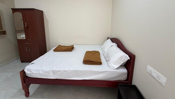 Een enkel bed met een witte matras en bruin beddengoed, een houten kledingkast en een witte muur met een stopcontact.