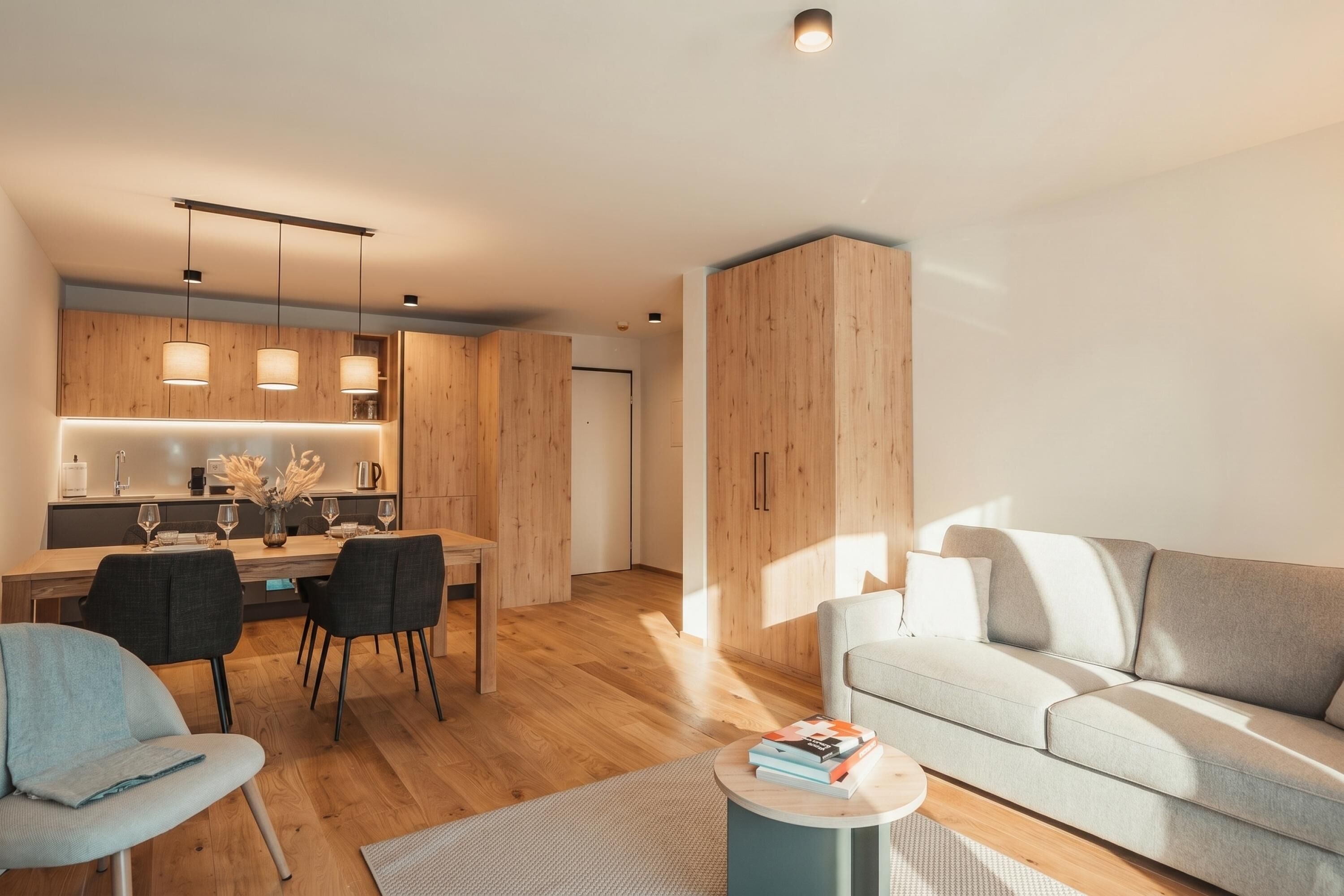 A modern living room with a sofa, a dining area with chairs and a table, and a kitchen with wooden cabinets and pendant lights.