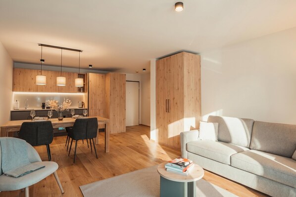 A modern living room with a sofa, a dining area with chairs and a table, and a kitchen with wooden cabinets and pendant lights.