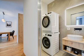 A modern laundry room with stacked washers and dryers, a wooden dining area, and a bathroom with a round sink. - Residenza Castanea 003 San Bernardino Alpine Apartments, San Bernardino, Switzerland (San Bernardino)