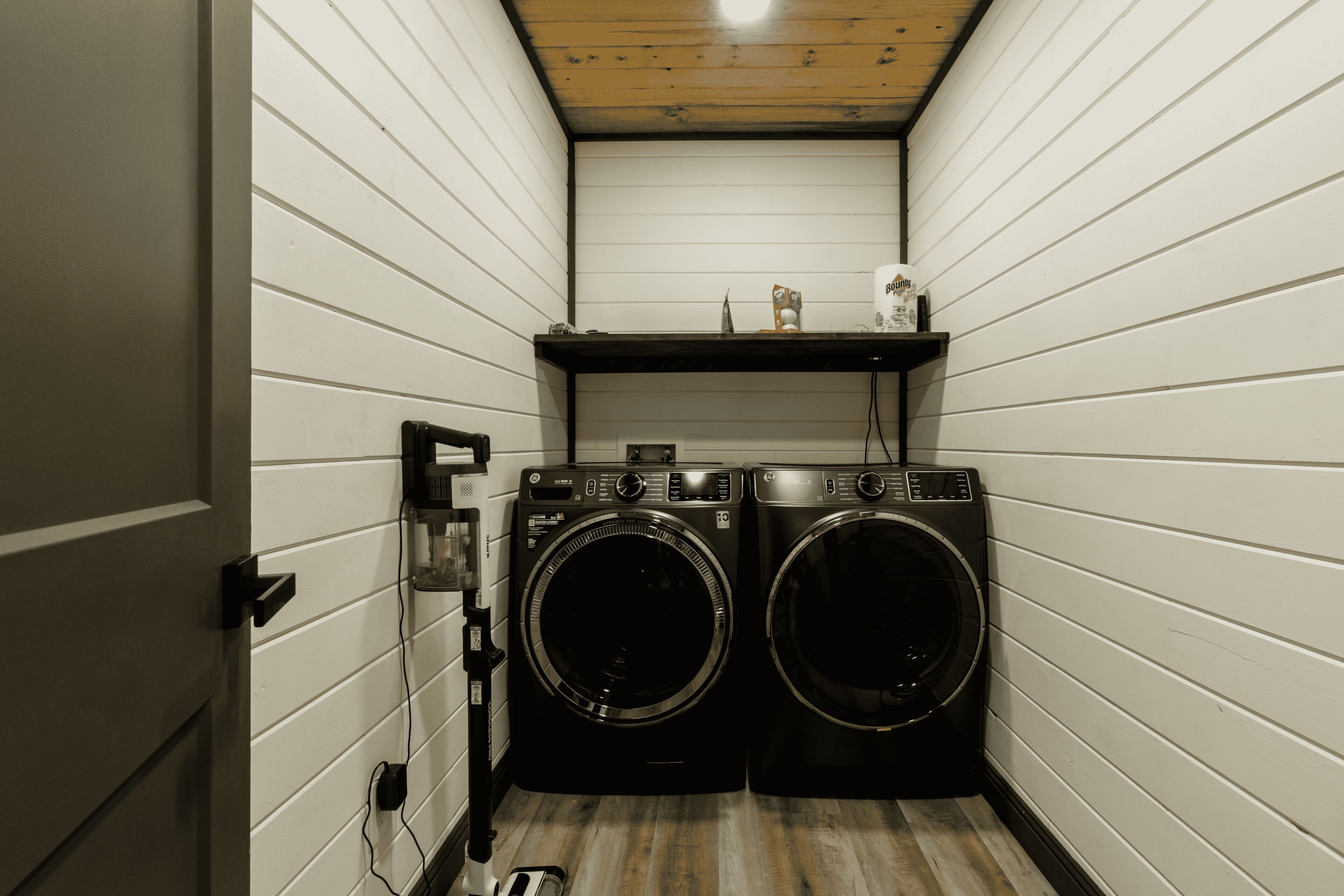 A laundry room with two front-loading washing machines, a shelf with items, and a wooden floor.