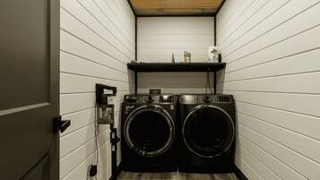 A laundry room with two front-loading washing machines, a shelf with items, and a wooden floor.
