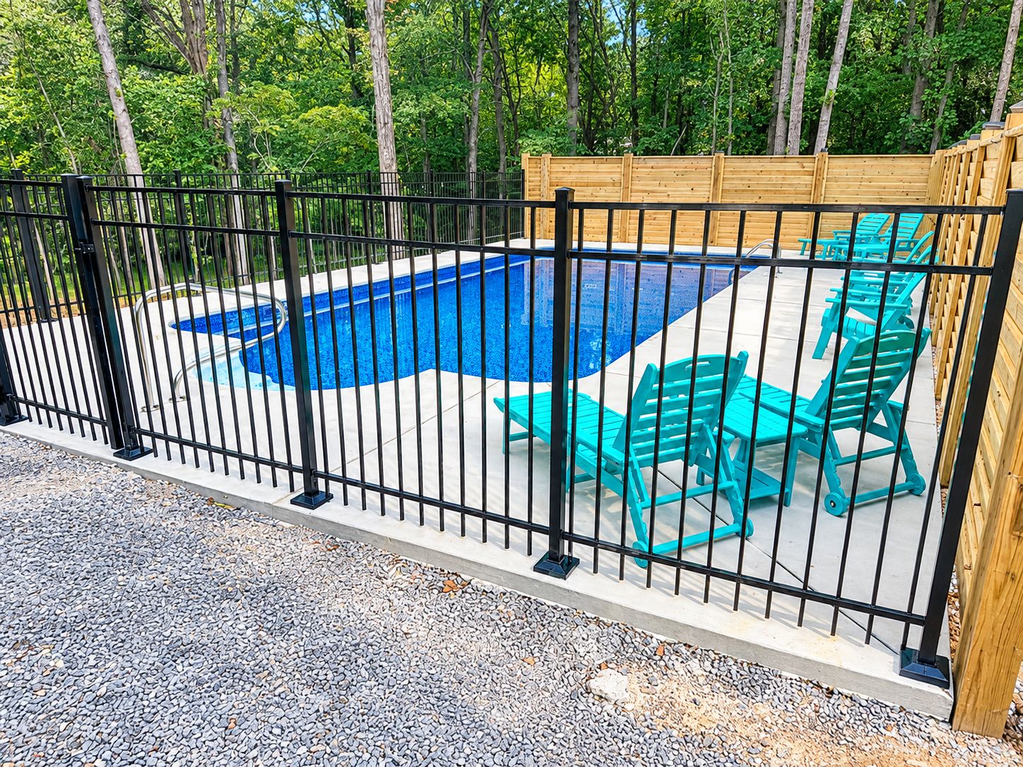 A swimming pool with a black metal fence, surrounded by gravel and trees.