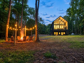 A cabin with a large glass front, surrounded by trees, and a fire pit with chairs.