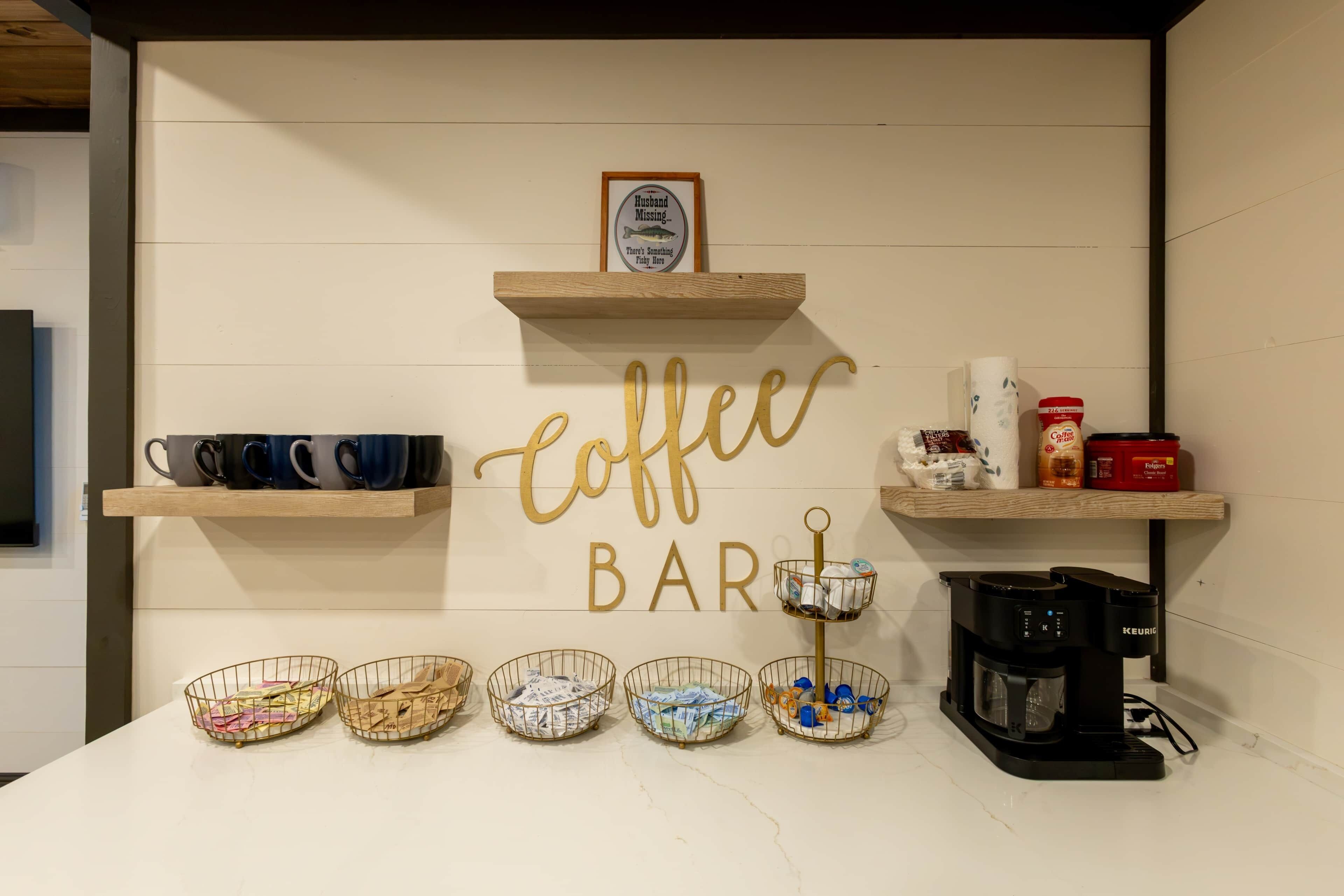 A coffee bar with a sign, coffee maker, mugs, and baskets of items on a counter.