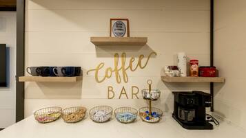 A coffee bar with a sign, coffee maker, mugs, and baskets of items on a counter.