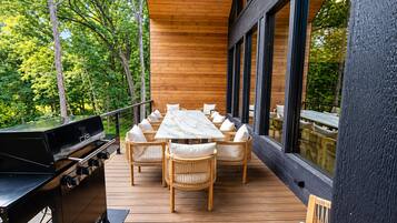 Une terrasse extérieure dotée d’un barbecue, de meubles en osier et de grandes fenêtres donnant sur une forêt.