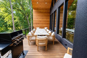 Una terrassa a l'exterior amb una barbacoa, mobiliari de vimet i grans finestres amb vista a un bosc.