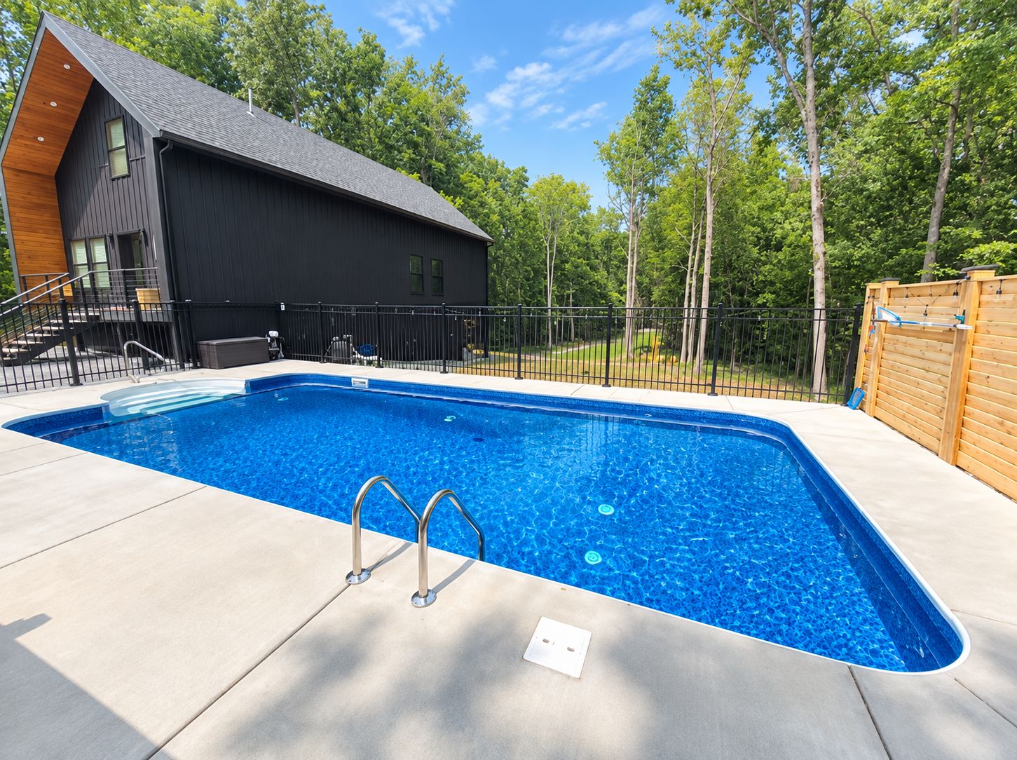 A rectangular swimming pool with a black fence, a wooden deck, and a modern house with a sloped roof.