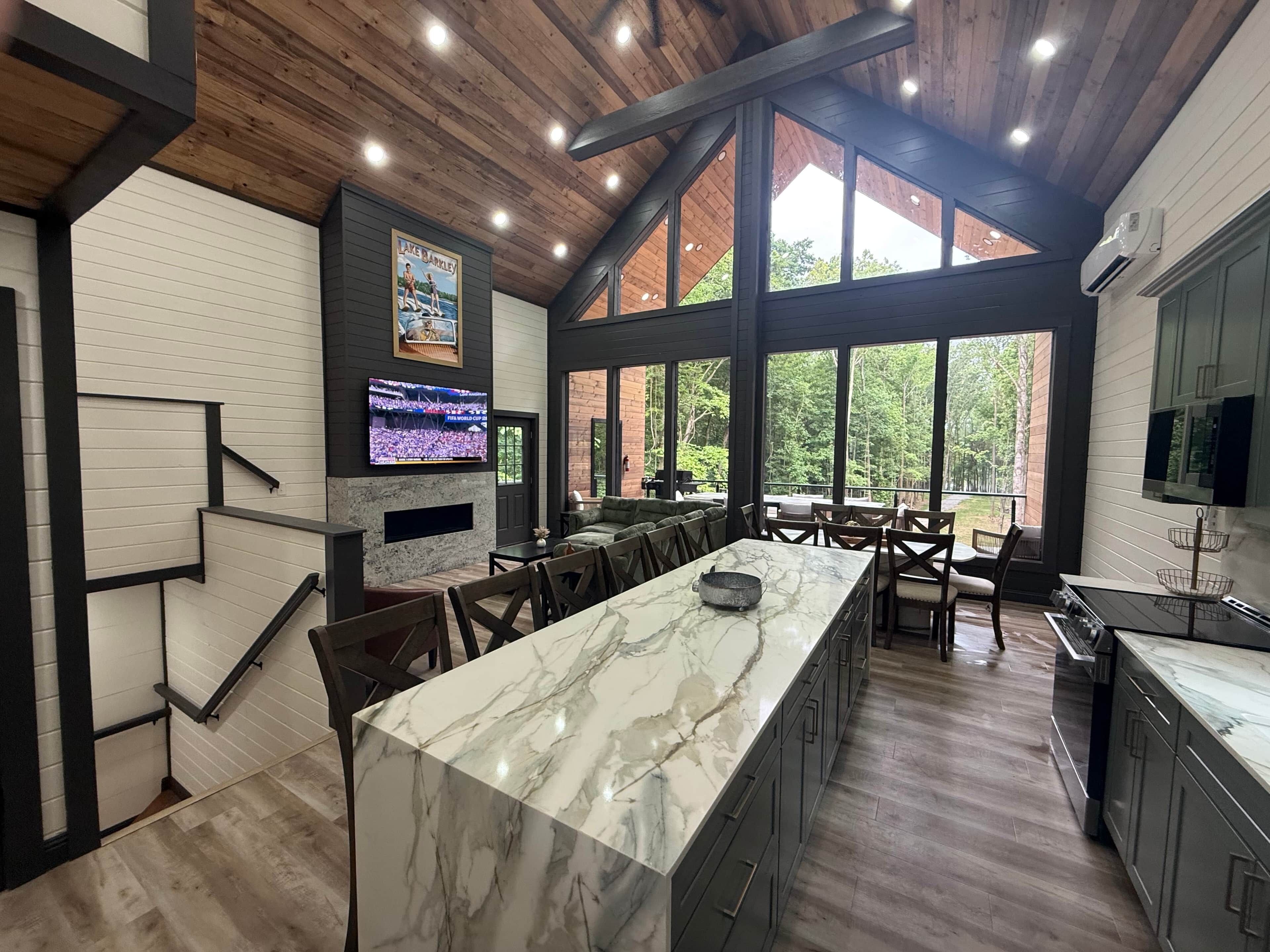 A spacious kitchen with a marble island, large windows, and a TV displaying a sports event.