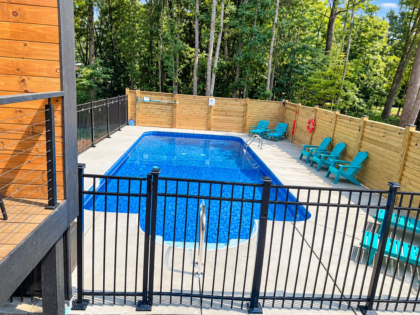 Piscina rectangular con cerca de metal negro, rodeada de una terraza de madera y sillas.