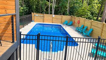Piscina rectangular con cerca de metal negro, rodeada de una terraza de madera y sillas.
