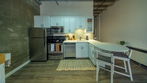 Cocina compacta con armarios blancos, un refrigerador de acero inoxidable, un microondas y una estufa. Cuenta con una mesa pequeña y una silla, y un tapete de cuadros en el suelo.