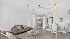 Ein modernes Wohnzimmer mit einer braunen Couch, einem runden Couchtisch mit Gebäck und einem Essbereich mit weißen Stühlen.