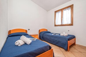 Ein Zimmer mit zwei Betten, jeweils mit einer blauen Bettdecke und einem orangefarbenen Kopfteil. Ein Fenster mit Holzrahmen und weißen Vorhängen ist ebenfalls vorhanden.