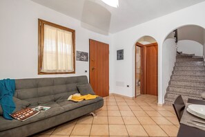 Ein Wohnzimmer mit grauem Sofa, einem Fenster mit leichtem Vorhang, einer Holztür und einer Treppe, die zu einer anderen Ebene führt.