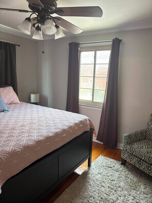 Une chambre à coucher avec un lit, un ventilateur de plafond, une fenêtre avec des rideaux, une chaise et une lampe.