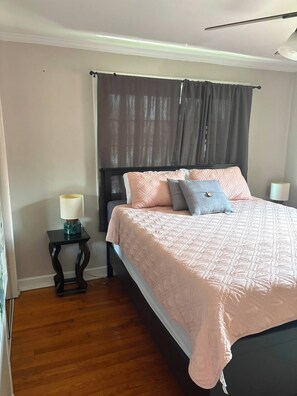 A bedroom with a bed, a bedside table with a lamp, a window with curtains, and a wooden floor. - 2BR Condo by Airport and Downtown Cbus (Columbus)
