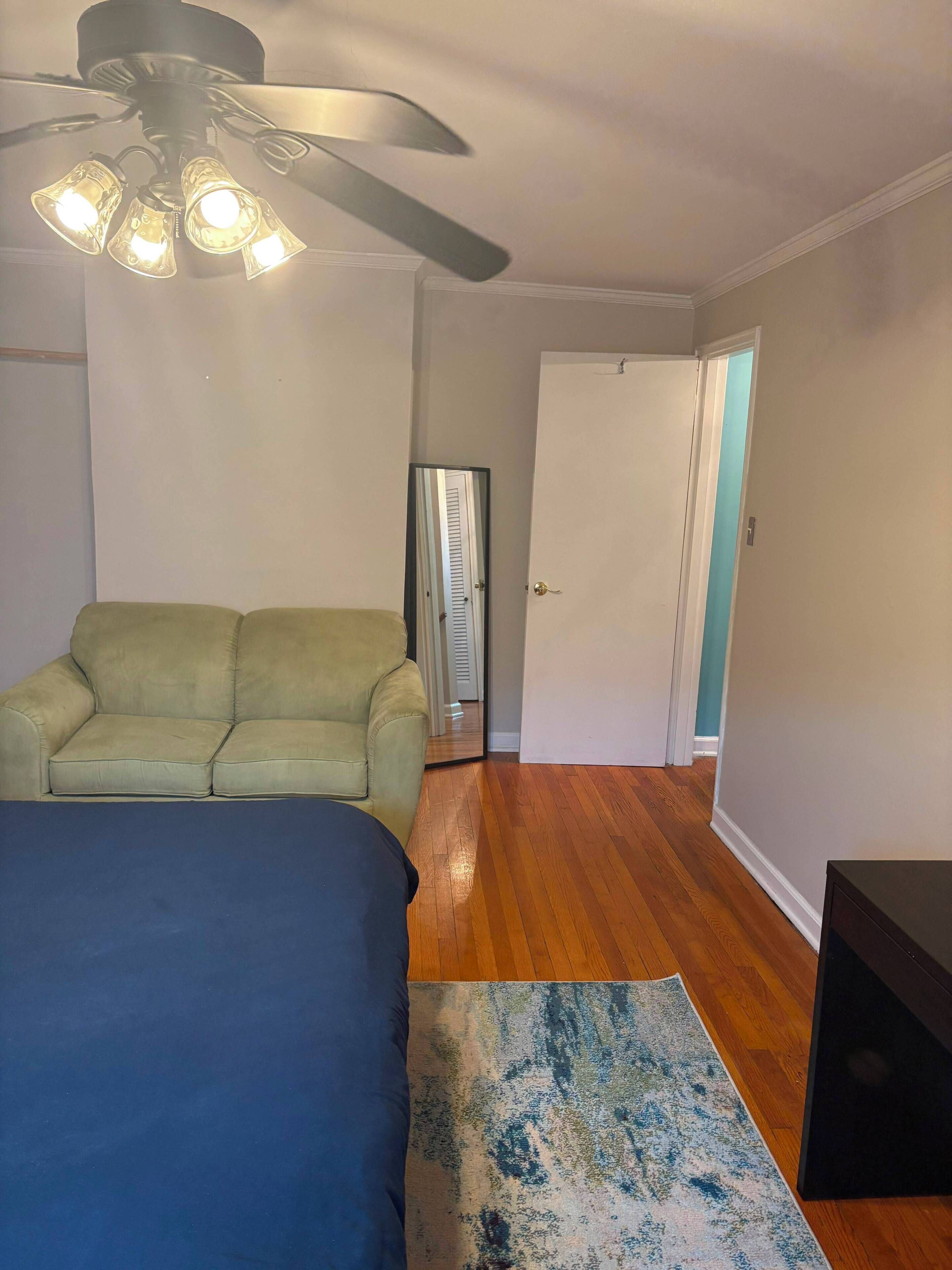 A bedroom with a bed, a green sofa, a ceiling fan, and a door leading to another room.