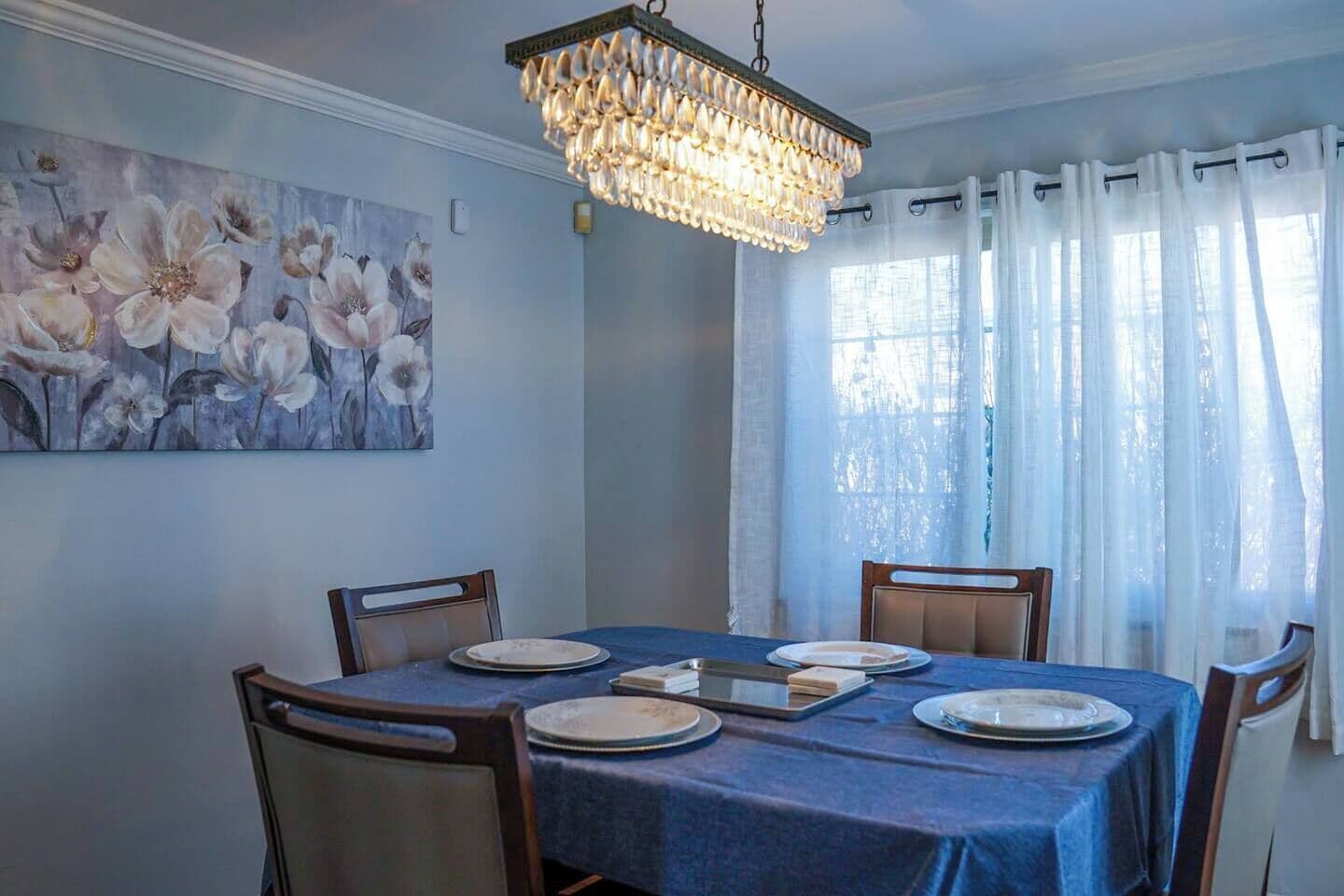 A dining room with a table set for four, a chandelier, a floral painting on the wall, and a window with sheer curtains.