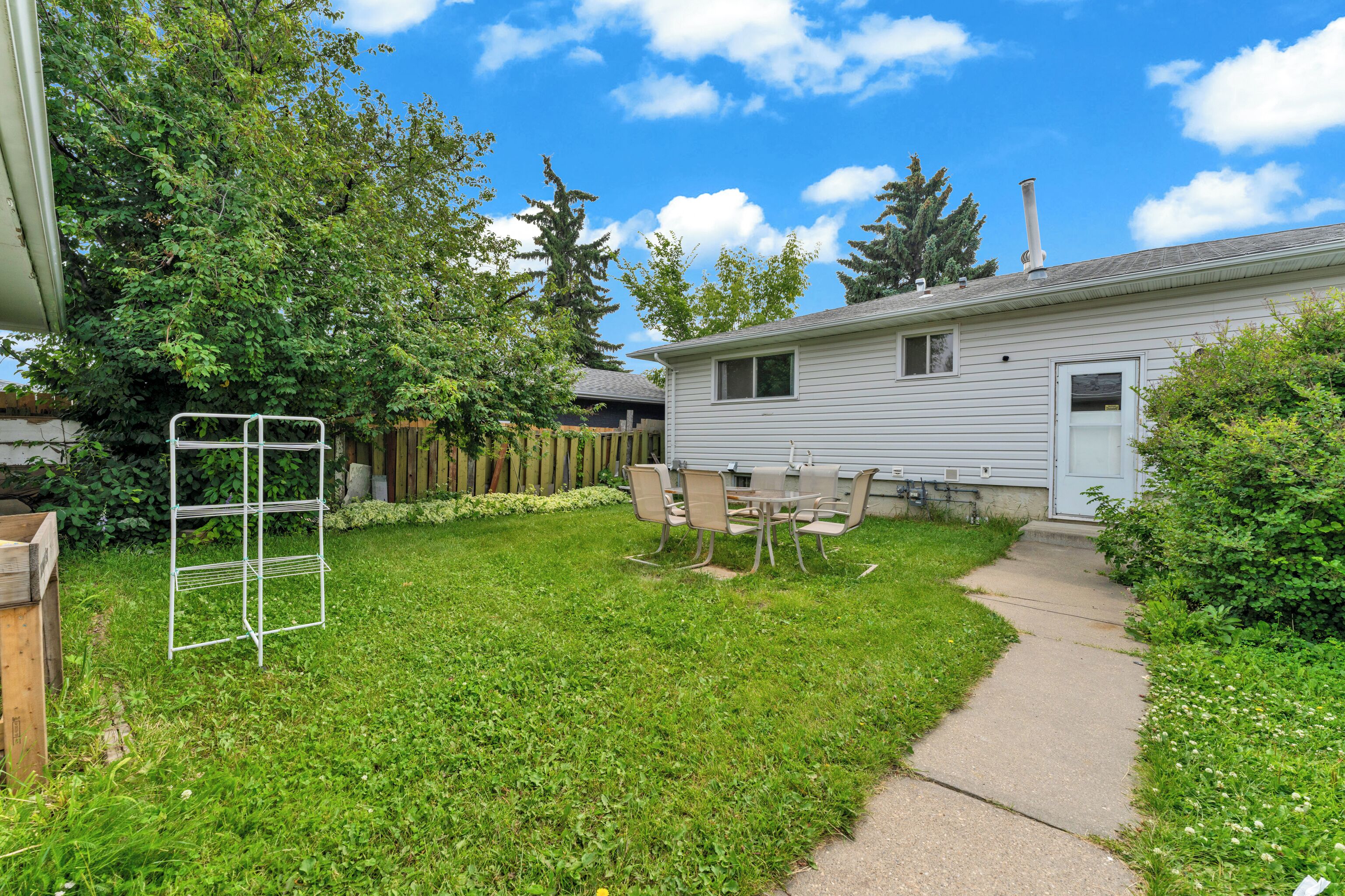 A residential backyard with a lawn, a metal chair and table set, a wooden fence, and a house with a chimney.