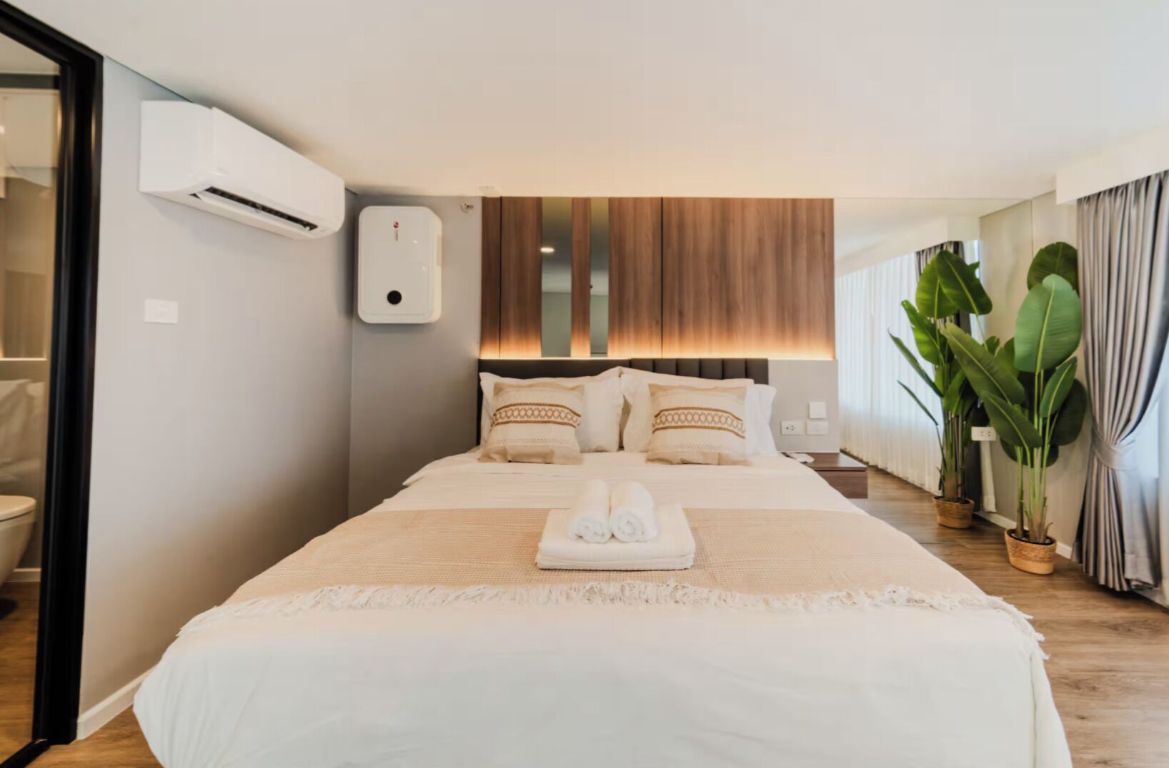 A hotel room with a large bed, a bedside table, an air conditioning unit, a wooden headboard, and a potted plant.