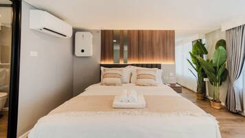 A hotel room with a large bed, a bedside table, an air conditioning unit, a wooden headboard, and a potted plant.