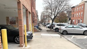 En parkeringsplads med biler parkeret langs en murstensbygning, et fortov og et træ i baggrunden.