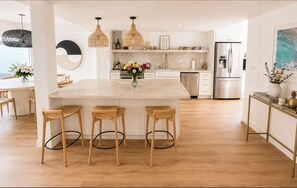 Et moderne køkken med en central ø, barstole, hængende lamper og et rustfrit stål køleskab.