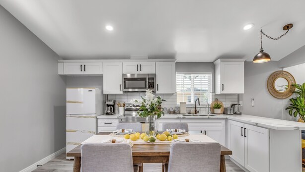 A modern kitchen with white cabinetry, a central island, and a dining area with two chairs. - Cozy Blue Cottage Near Old Town Monrovia, City of Hope. Pet-Friendly (Monrovia)