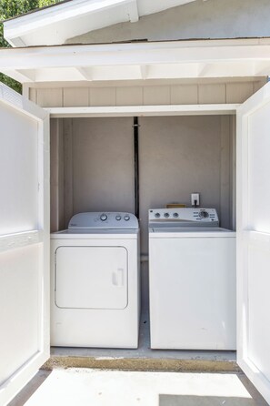 Een wasserette met twee witte wasmachines en een droger, omgeven door een witte buitenmuur en een schuin dak.