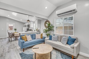 Een moderne woonkamer met een blauwe bank, een beige bank en een ronde salontafel. Er is een eethoek met een tafel en stoelen, en een keuken op de achtergrond.