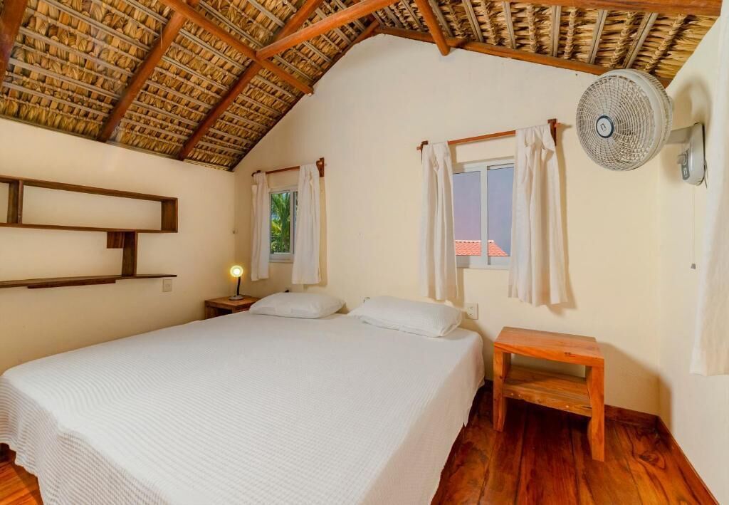 A bedroom with a bed, a nightstand, a wooden bench, a ceiling fan, and a window with curtains.