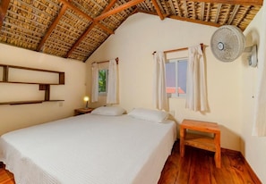 A bedroom with a bed, a nightstand, a wooden bench, a ceiling fan, and a window with curtains. - Cabaña Paraguitos (Brisas de Zicatela)