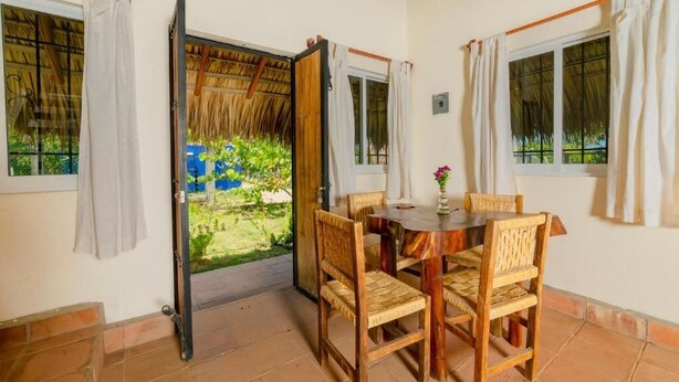 A dining area with a table and chairs, a view of the garden through an open door, and a window with a curtain. - Cabaña Paraguitos (Brisas de Zicatela)
