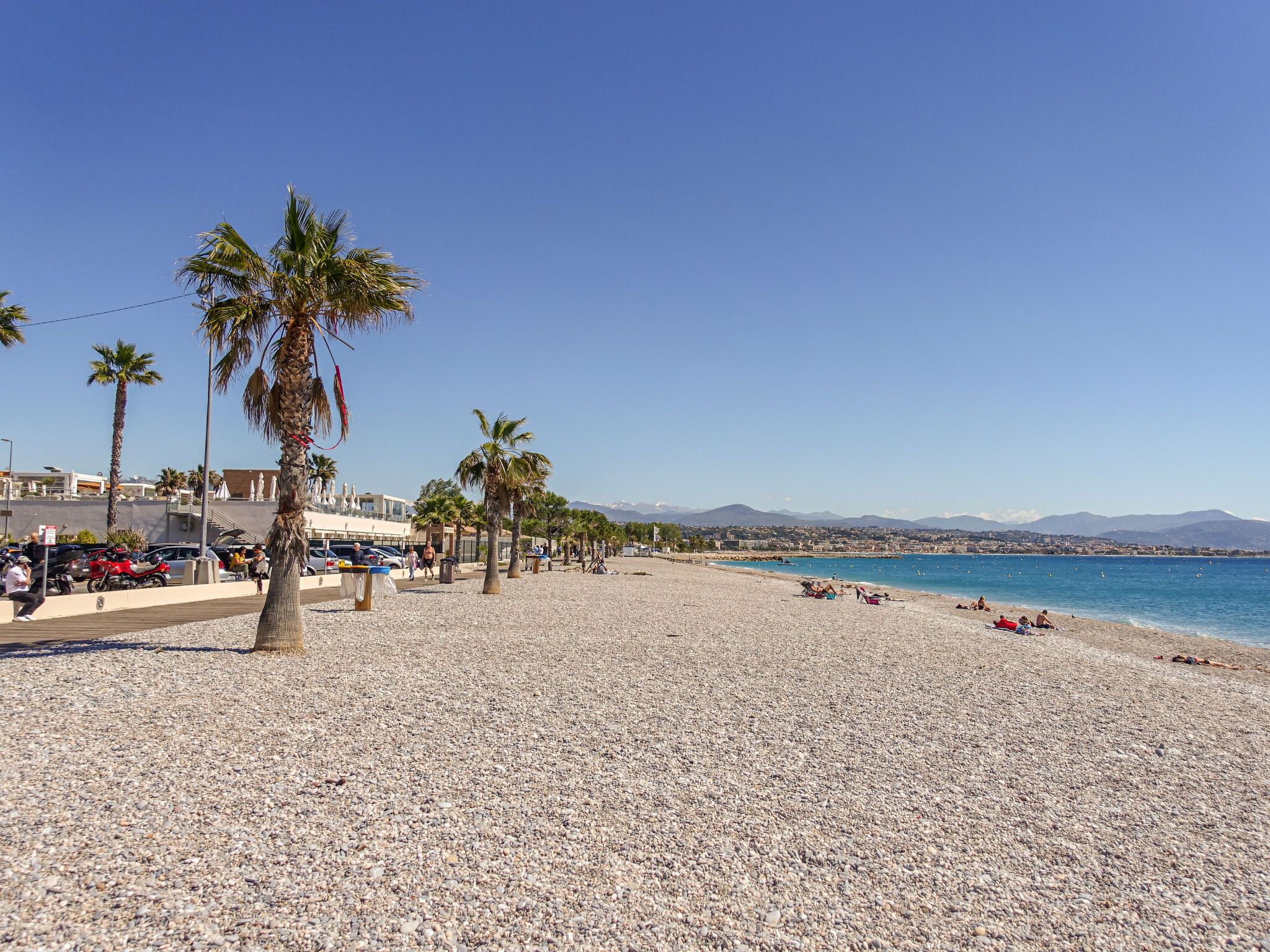 Ein Kiesstrand mit Palmen, blauem Himmel und einem fernen Gebirgszug.