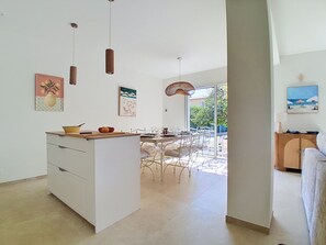 A modern kitchen with white cabinetry, a dining area with a large table and chairs, and artwork on the walls. - Villa l'Orangeraie by Interhome (Villeneuve-Loubet)