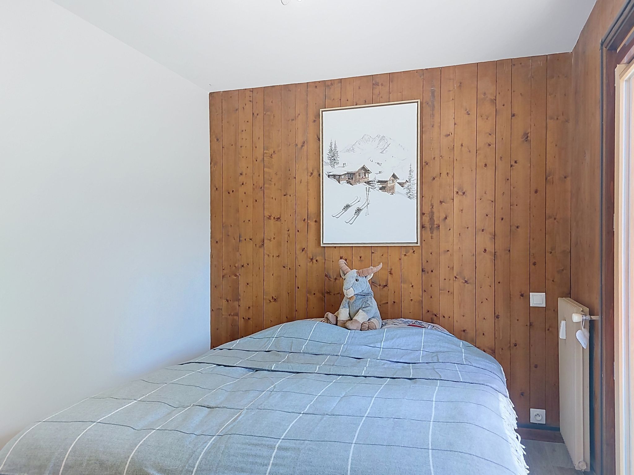 Une chambre avec un mur lambrissé en bois, un lit recouvert d’une courtepointe bleue et un tableau encadré au-dessus du lit.