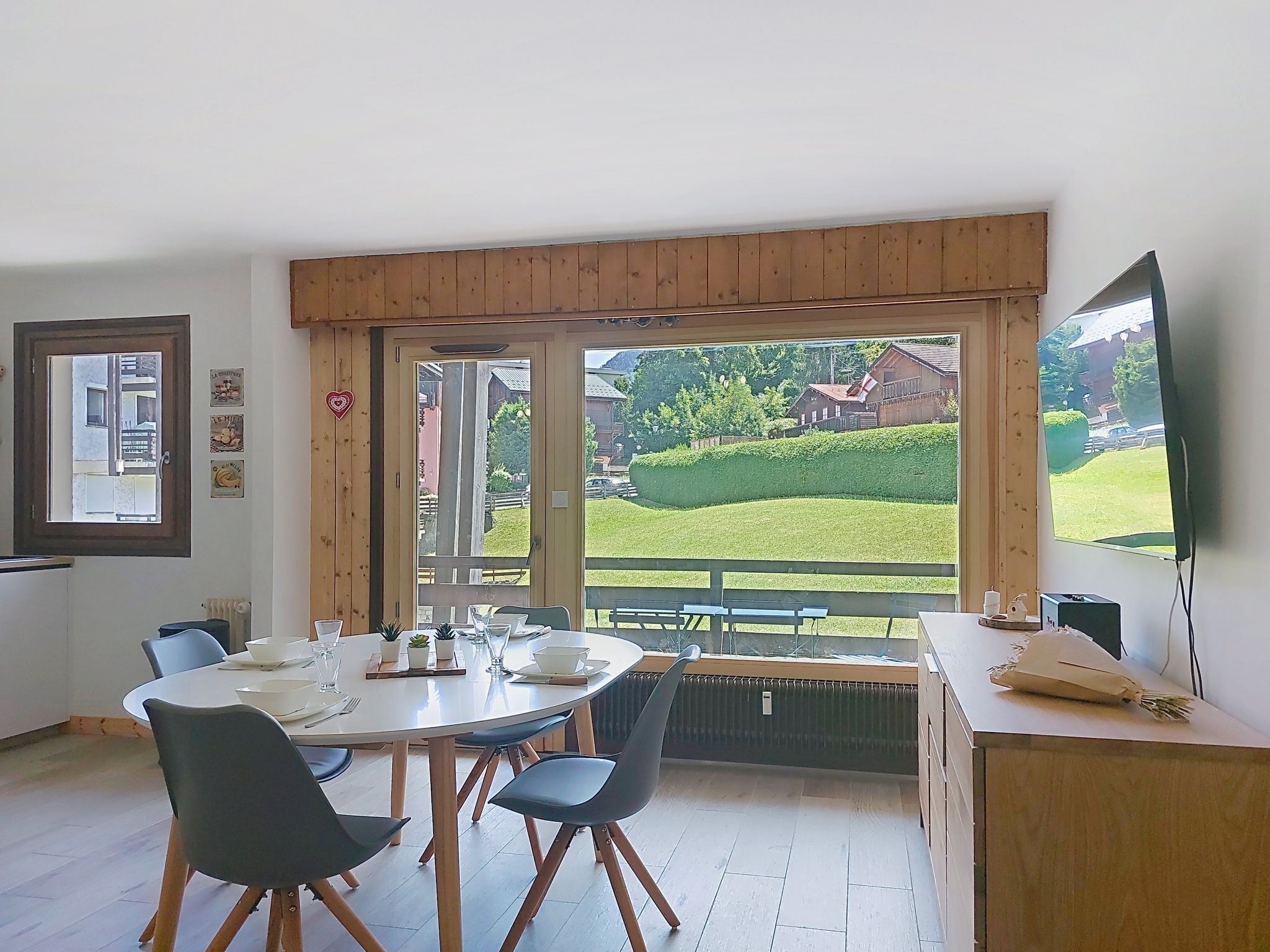 A dining area with a table set for four, wooden beams above, and a view of a green landscape through large windows.