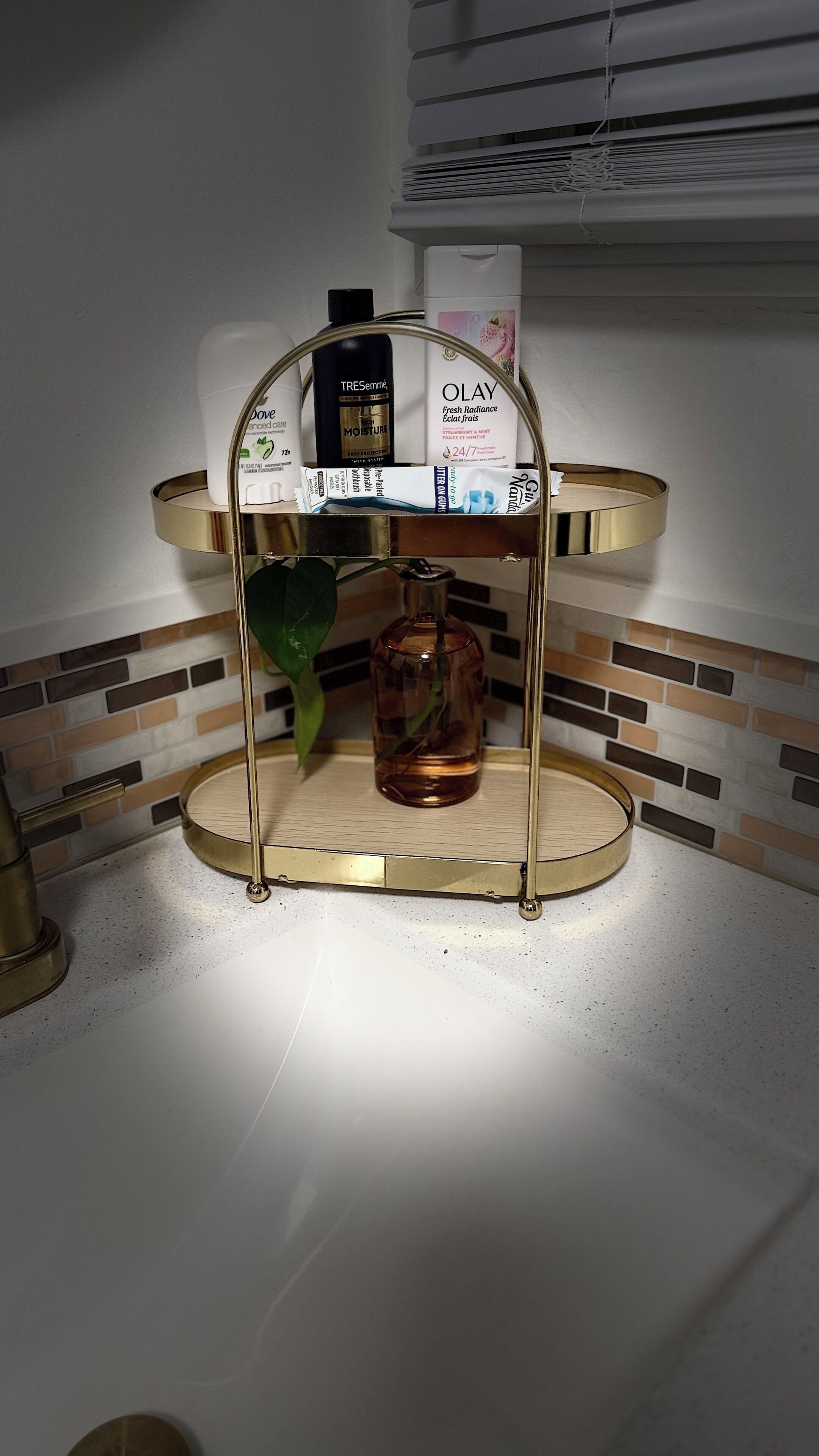 Ein Badezimmer-Waschtisch mit einem goldfarbenen Spiegelrahmen, einer Dove Body Lotion Flasche, einer Olay Feuchtigkeitscreme Flasche und einer Tressemé Körperöl Flasche.