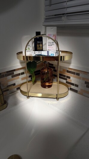 Ein Badezimmer-Waschtisch mit einem goldfarbenen Spiegelrahmen, einer Dove Body Lotion Flasche, einer Olay Feuchtigkeitscreme Flasche und einer Tressemé Körperöl Flasche.