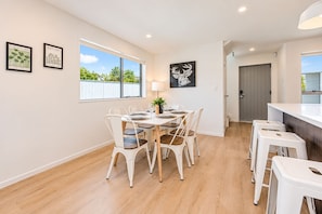 A dining area with a table set for four, white chairs, a lamp, and framed artwork on the walls. - Modern Central Auckland Stay- perfect for families, groups & business travellers (Mt Roskill)