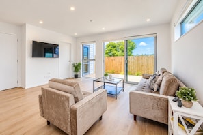 A modern living room with a flat-screen TV, two sofas, a coffee table, and a sliding glass door leading to a backyard. - Modern Central Auckland Stay- perfect for families, groups & business travellers (Mt Roskill)