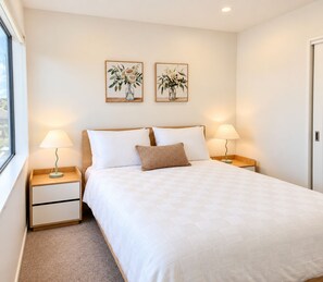 A neatly made bed with two pillows and a single decorative pillow, flanked by bedside tables with lamps, in a room with a plain wall and a door. - Modern Central Auckland Stay- perfect for families, groups & business travellers (Mt Roskill)