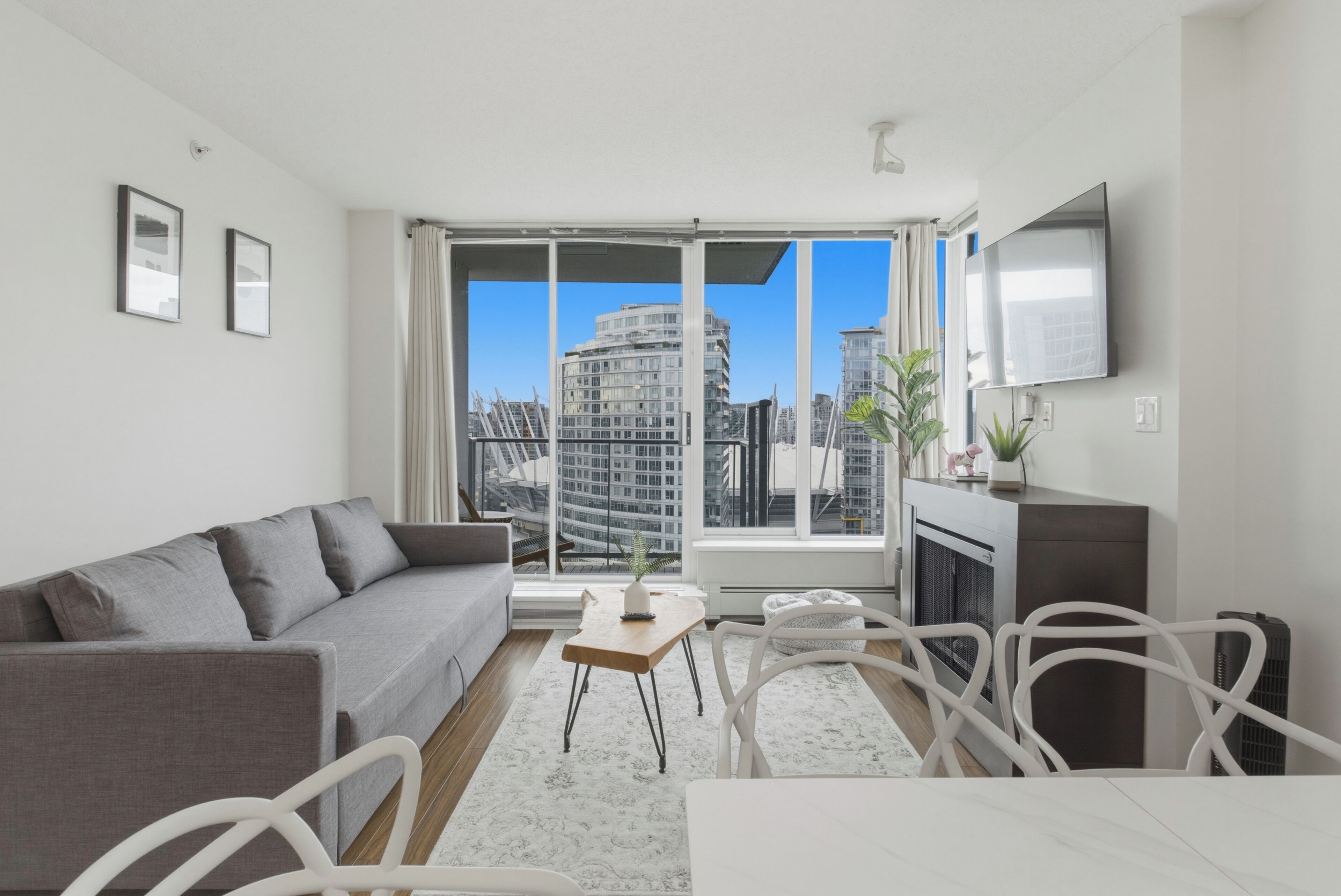 Ein modernes Wohnzimmer mit grauem Sofa, einem kleinen Tisch, einem Fernseher und Blick auf Stadtgebäude durch große Fenster.
