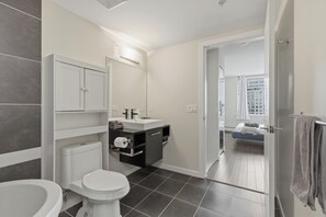 Een moderne badkamer met toilet, wastafel en spiegel. Een deur is zichtbaar, die leidt naar een slaapkamer met een bed en raam.