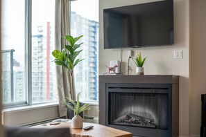 Een woonkamer met een open haard, een flatscreen-televisie die erboven is bevestigd, een raam met uitzicht op gebouwen, en een plant in een pot op de vensterbank.